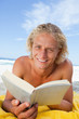 Smiling blonde man reading a book while lying on the beach