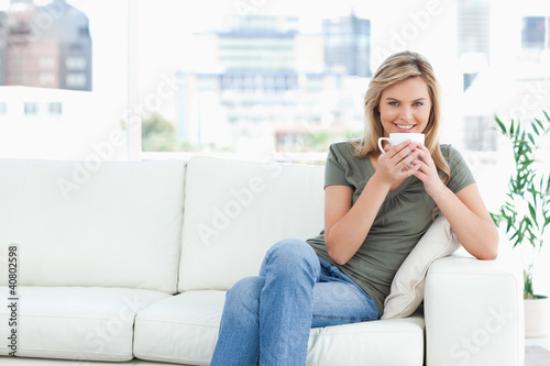 Photo: Woman smiling with a cup near her lips, sitting on the couch and Woman smiling with a cup near her lips, sitting on the couch and