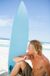 Serious blonde man sitting on the beach with his surfboard next