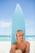 Serious blonde man sitting down in front of his surfboard