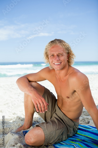 Smiling man sitting on his beach towel while looking at the came