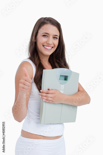 Foto: Smiling brunette putting her thumbs up while holding weighing sc Smiling brunette putting her thumbs up while holding weighing sc