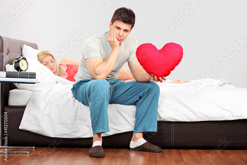 Photo: Man in thoughts holding a heart shaped pillow while his girlfrie Man in thoughts holding a heart shaped pillow while his girlfrie