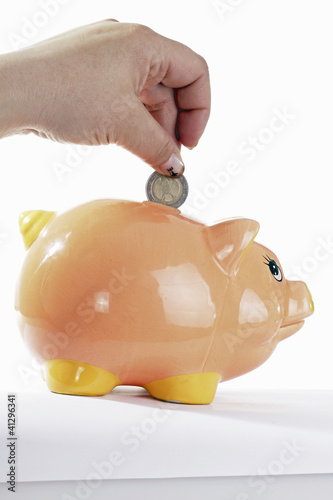 Photo: piggy bank with hand with euro coin on rhe white background piggy bank with hand with euro coin on rhe white background