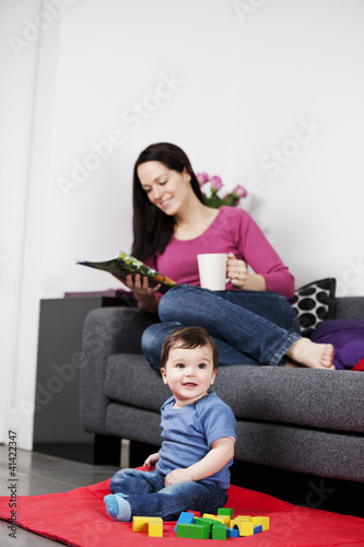 Photo: A mother relaxing on a sofa with a magazine, baby playing A mother relaxing on a sofa with a magazine, baby playing