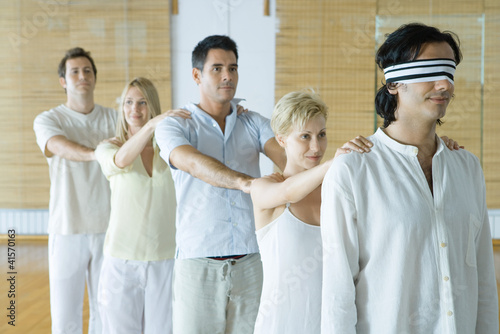 Photo: Group of adults standing single file holding onto each others' shoulders, leader blindfolded Group of adults standing single file holding onto each others' shoulders, leader blindfolded