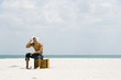 Photo: Man sitting on suitcase at the beach, covering head with handkerchief, looking up at sun Man sitting on suitcase at the beach, covering head with handkerchief, looking up at sun