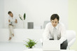 Photo: Professional man using laptop computer in casual office, female colleague reading in background Professional man using laptop computer in casual office, female colleague reading in background
