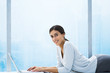 Woman lying down in front of bay window, using laptop, smiling at camera