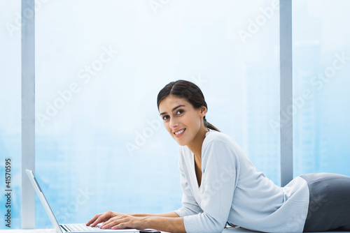Photo: Woman lying down in front of bay window, using laptop, smiling at camera Woman lying down in front of bay window, using laptop, smiling at camera