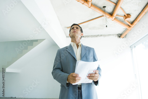 Photo: Well-dressed man holding blueprints, inspecting unfinished home interior Well-dressed man holding blueprints, inspecting unfinished home interior
