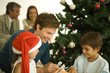 Father and two children sitting by Christmas tree, opening presents together, daughter wearing Santa hat