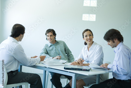 Photo: Business associates seated at table, chatting, one smiling at camera Business associates seated at table, chatting, one smiling at camera