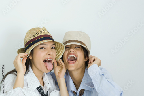 Photo: Two young friends wearing hats, sticking out tongues, looking at camera, portrait Two young friends wearing hats, sticking out tongues, looking at camera, portrait