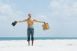 Man standing at the beach, holding up shoes and suitcase, head back