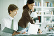 Photo: Male and female colleagues at desk, man looking at laptop computer, woman using phone Male and female colleagues at desk, man looking at laptop computer, woman using phone
