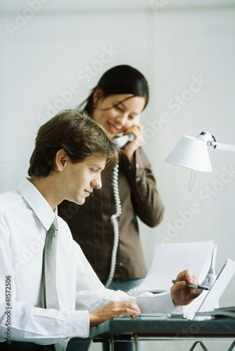 Photo: Male and female colleagues looking at laptop computer together, man pointing with pen, woman holding phone Male and female colleagues looking at laptop computer together, man pointing with pen, woman holding phone