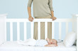 Mother standing behind crib as infant sleeps, cropped view