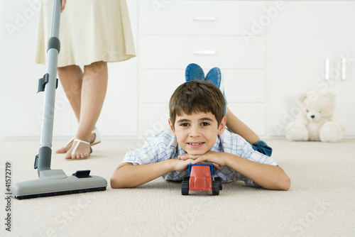 Photo: Little boy lying on the ground with toys, smiling at camera, mother vacuuming around him Little boy lying on the ground with toys, smiling at camera, mother vacuuming around him