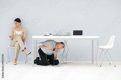 Photo: Man kneeling under desk, connecting tangled wires, woman sitting in chair, looking at hand Man kneeling under desk, connecting tangled wires, woman sitting in chair, looking at hand