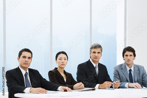 Photo: Business associates sitting at table, looking at camera, serious expressions Business associates sitting at table, looking at camera, serious expressions