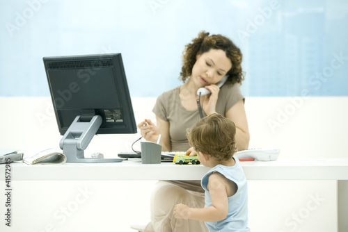 Photo: Woman using phone in office, toddler son playing with toy truck on desk Woman using phone in office, toddler son playing with toy truck on desk