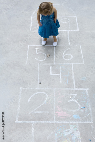 Photo: Little girl standing on hopscotch grid, looking down at rock, high angle view Little girl standing on hopscotch grid, looking down at rock, high angle view