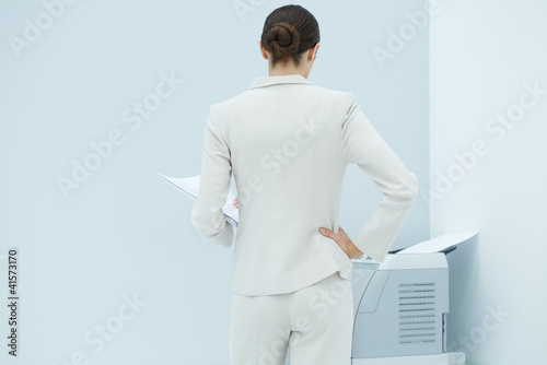 Photo: Businesswoman standing with hand on hip, looking at printer, rear view Businesswoman standing with hand on hip, looking at printer, rear view