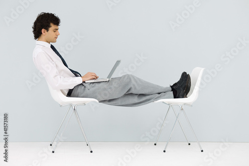 Photo: Businessman sitting in chair, feet up on another chair, using laptop Businessman sitting in chair, feet up on another chair, using laptop