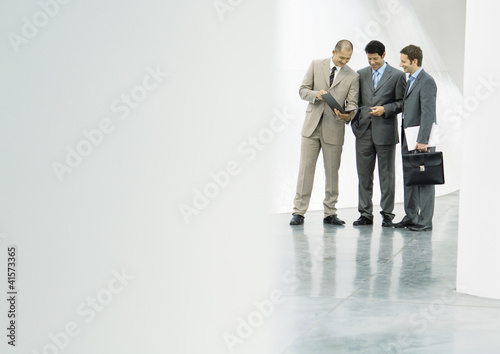 Photo: Three businessman standing in lobby, looking at file Three businessman standing in lobby, looking at file