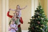 Photo: Family of four by Christmas tree, girl (4-6) with angel decoration on father's shoulders, portrait. Family of four by Christmas tree, girl (4-6) with angel decoration on father's shoulders, portrait.