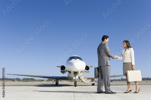 Photo: Businessman and woman with briefcase shaking hands by aeroplane on runway, low angle view Businessman and woman with briefcase shaking hands by aeroplane on runway, low angle view