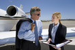 Photo: Businessman and woman looking at folder by aeroplane on runway, smiling, close-up Businessman and woman looking at folder by aeroplane on runway, smiling, close-up
