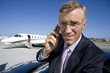 Businessman using mobile phone by car, on runway by aeroplane, smiling, portrait