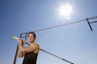 Photo: Young male pole vault athlete with pole by bar, portrait, low angle view (sun flare) Young male pole vault athlete with pole by bar, portrait, low angle view (sun flare)