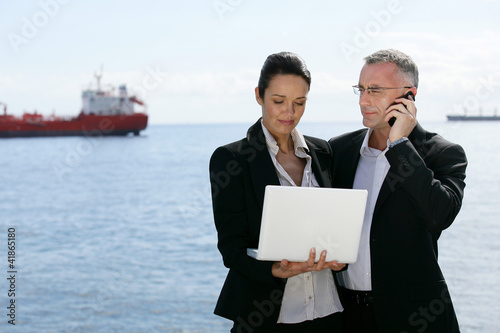 Photo: Two businesspeople stood with a laptop by the coast Two businesspeople stood with a laptop by the coast