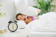 Upset woman closing ears by pillow to avoid hearing alarm clock