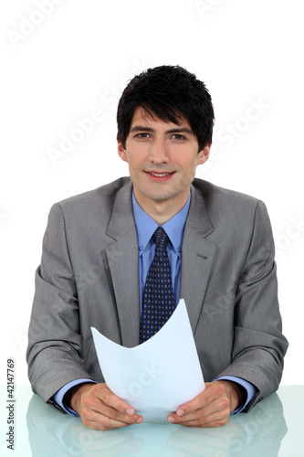 Photo: businessman sitting at his desk holding a sheet of paper businessman sitting at his desk holding a sheet of paper