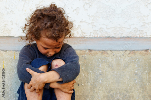 Photo: poor, sad little child girl sitting against the concrete wall poor, sad little child girl sitting against the concrete wall