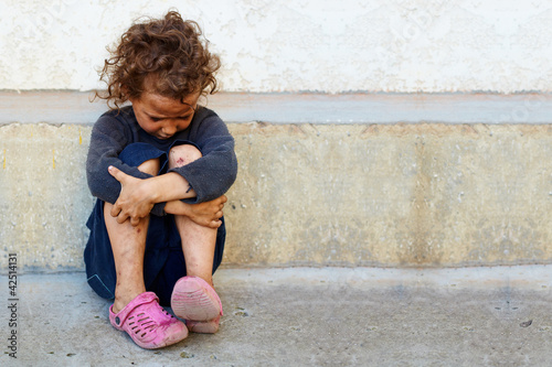 Photo: poor, sad little child girl sitting against the concrete wall poor, sad little child girl sitting against the concrete wall