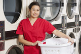 Photo: Portrait of a happy woman carrying laundry basket with washing machine in background Portrait of a happy woman carrying laundry basket with washing machine in background