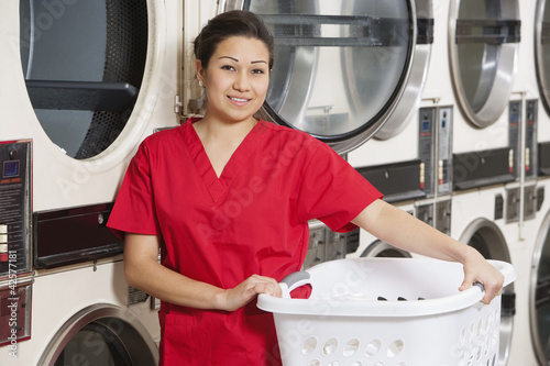 Photo: Portrait of a happy woman carrying laundry basket with washing machine in background Portrait of a happy woman carrying laundry basket with washing machine in background