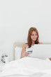 Photo: Close-up of a redheaded sitting on her bed while using her credit card online Close-up of a redheaded sitting on her bed while using her credit card online