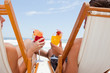 Photo: Rear view of a young couple holding their cocktails while sitting on deck chairs Rear view of a young couple holding their cocktails while sitting on deck chairs