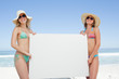 Two friends wearing sunglasses and hats while holding a blank poster