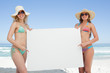 Two women in beach gear holding a blank poster by the sea
