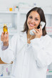 Smiling female pharmacist holding a bottle of pills while holding a phone