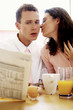 A man getting a kiss from his wife while having breakfast and reading newspaper