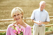 Foto: Senior woman holding a bouquet of flowers with her husband standing in the background Senior woman holding a bouquet of flowers with her husband standing in the background
