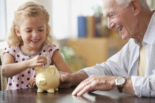 Photo: Caucasian grandfather watching granddaughter putting coin into piggy bank Caucasian grandfather watching granddaughter putting coin into piggy bank